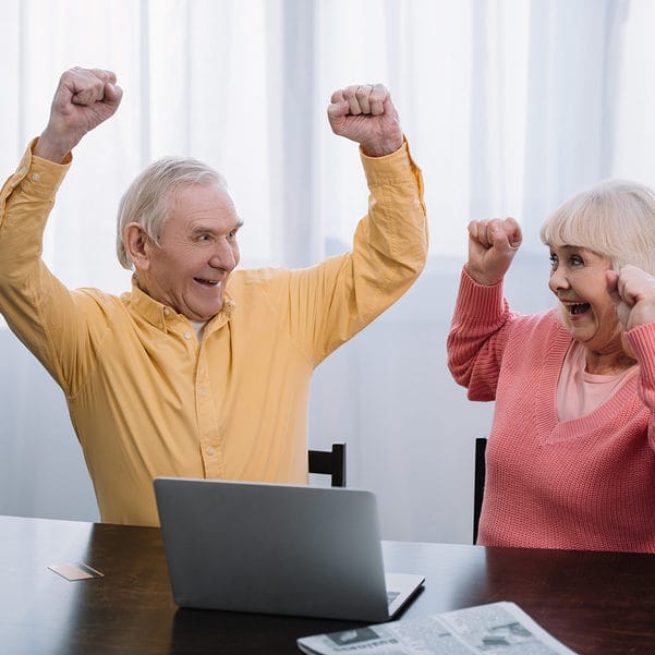 happy senior couple in colorful clothes sitting at table with laptop and cheering with hands in air